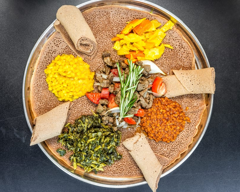 Ethiopian injera platter with various stews, vegetables, and meat, garnished with rosemary.