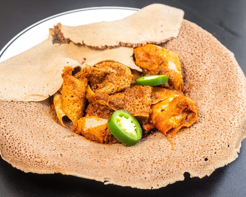Spicy Ethiopian Doro Wat chicken stew with injera bread and sliced jalapeños on a white plate.