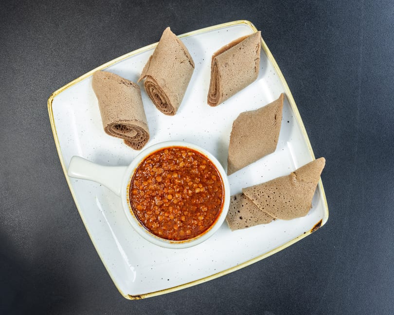 A white rectangular plate holds a bowl of red lentil stew surrounded by injera bread pieces.