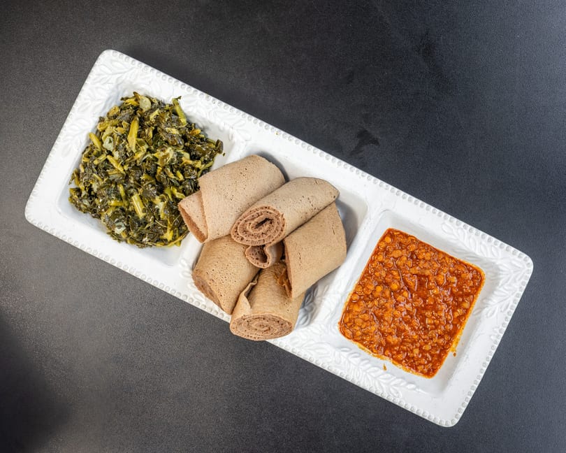 A white rectangular plate with three sections, holding green collard greens, rolled injera bread, and red lentils.