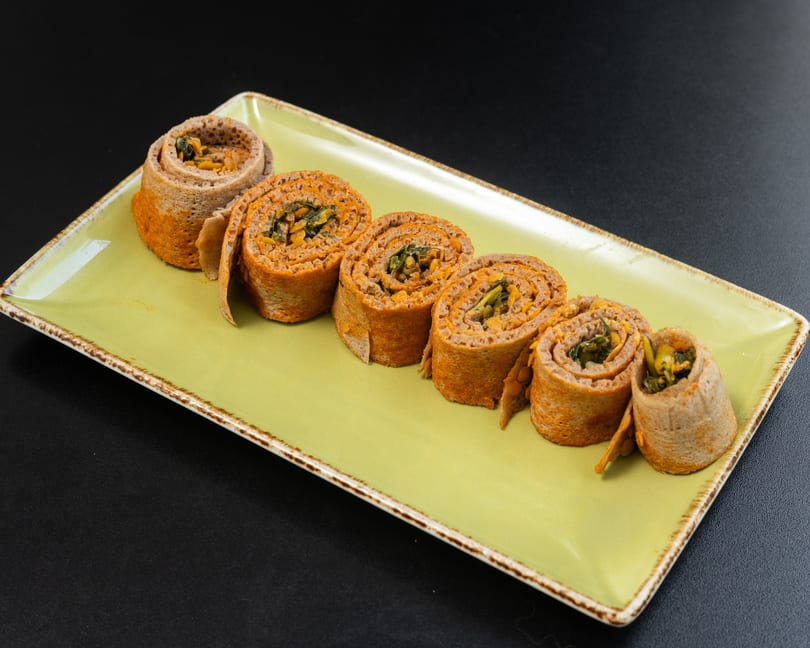 Six injera rolls filled with orange lentils and green vegetables, arranged on a light green rectangular plate.