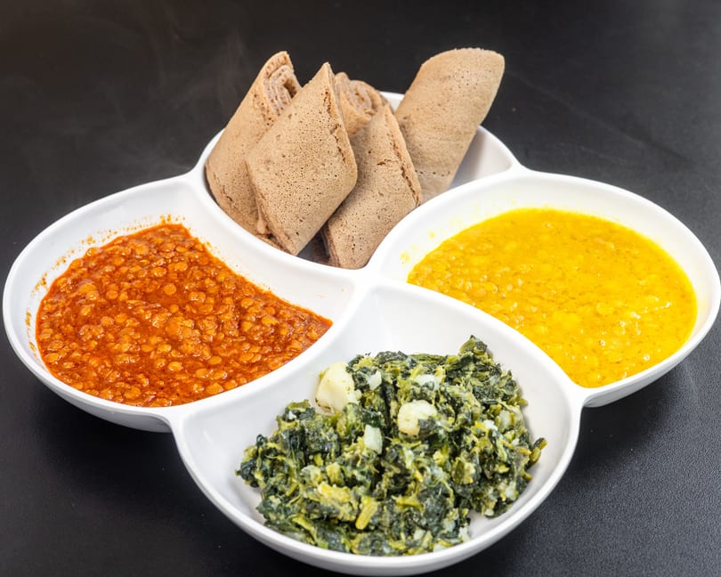 A white divided dish holds injera bread, red lentils, yellow split peas, and collard greens.