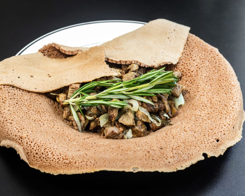 Ethiopian injera bread with meat, onions, and rosemary on a white plate.