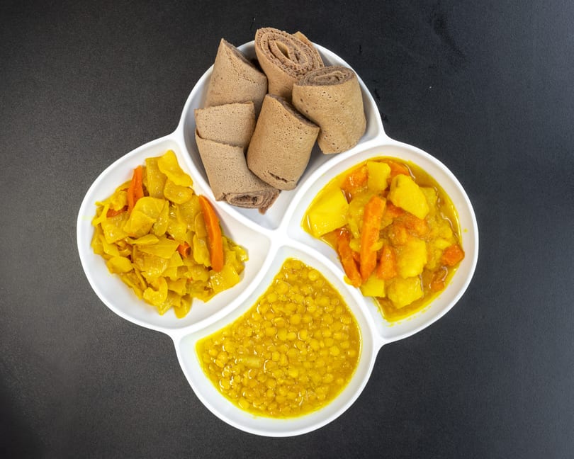A white clover-shaped dish with injera bread, yellow lentils, and two vegetable stews.