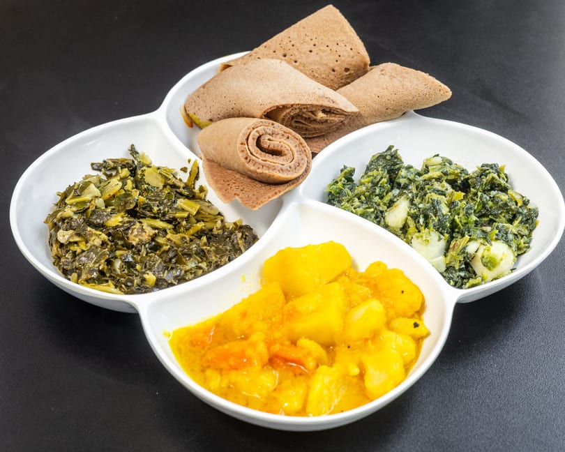A white segmented dish holds injera bread, collard greens, spinach, and potato stew on a dark surface.