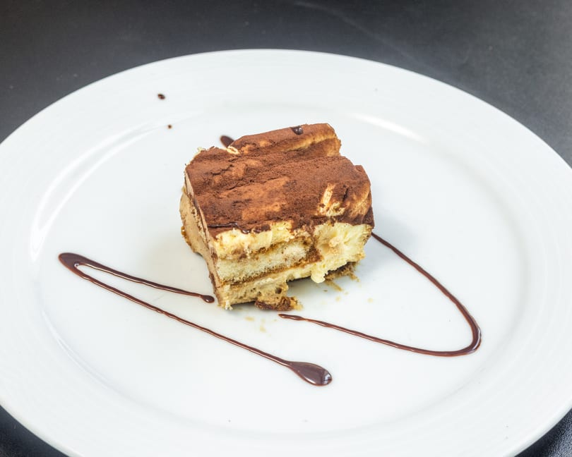 A slice of tiramisu with cocoa powder and chocolate sauce on a white plate.