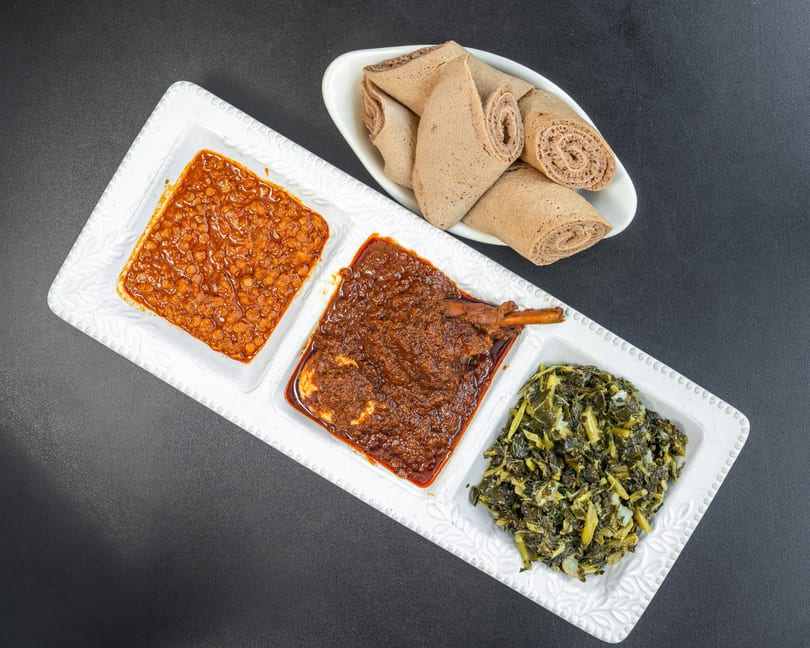 A white rectangular platter with three Ethiopian dishes: lentils, chicken stew, and collard greens, with injera bread.