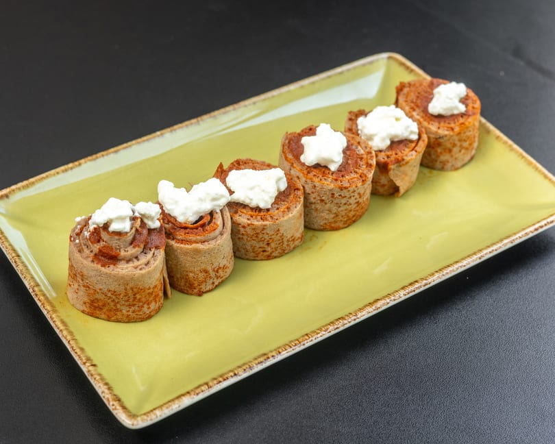 Five rolled crepes with a red filling and white cream topping, on a light green rectangular plate.