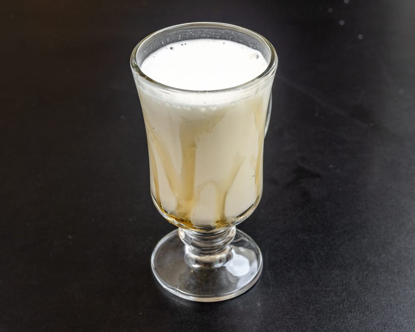 A tall glass of creamy white drink with honey drizzled down the sides, on a dark surface.