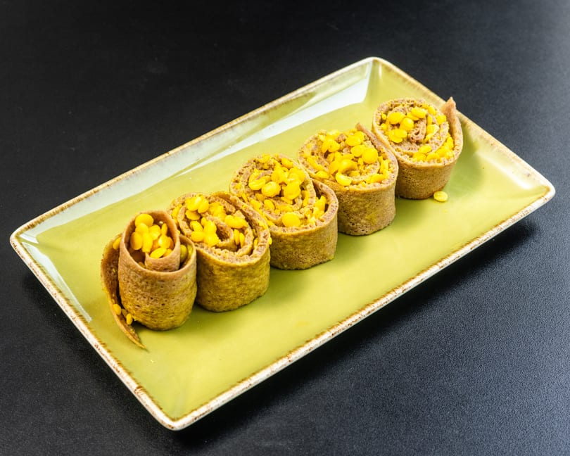 Five corn-filled crepes, rolled and arranged on a rectangular green plate against a dark background.